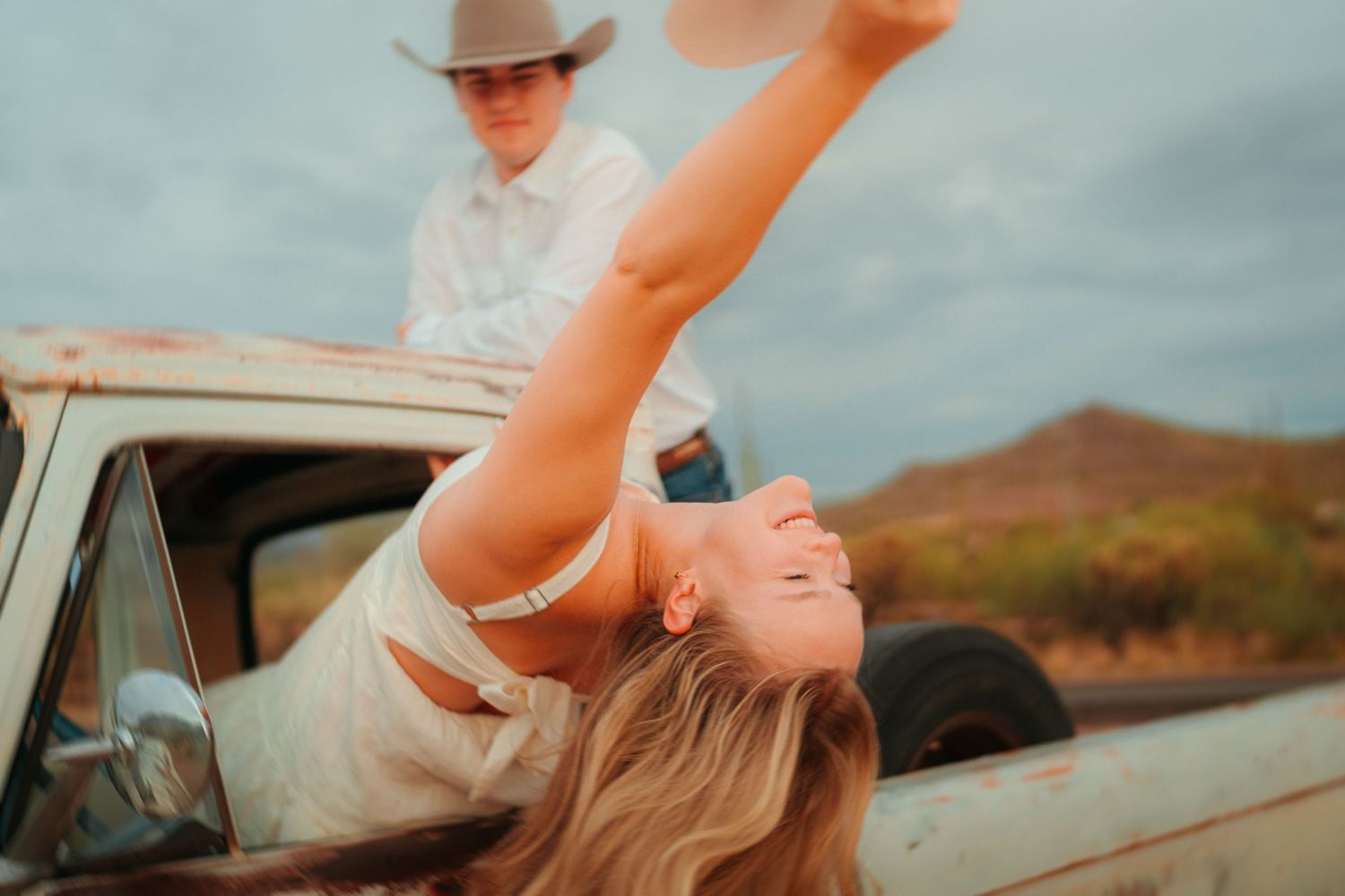 Woman leaning out of vintage truck window holding cowboy hat, with man standing in truck bed, desert landscape.