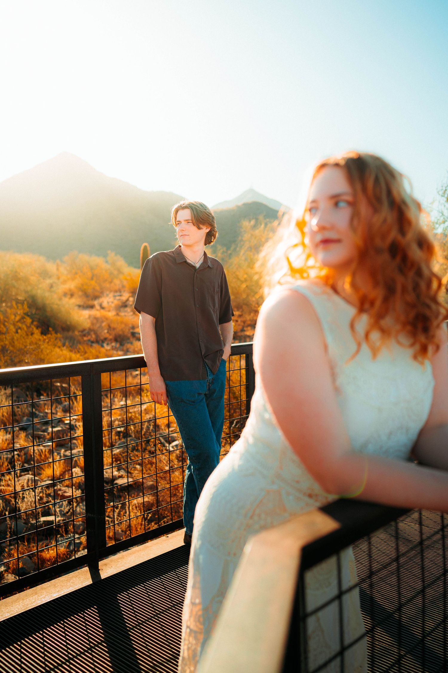 Redhead woman in white dress and man in black shirt posing on a railing with desert mountains behind.