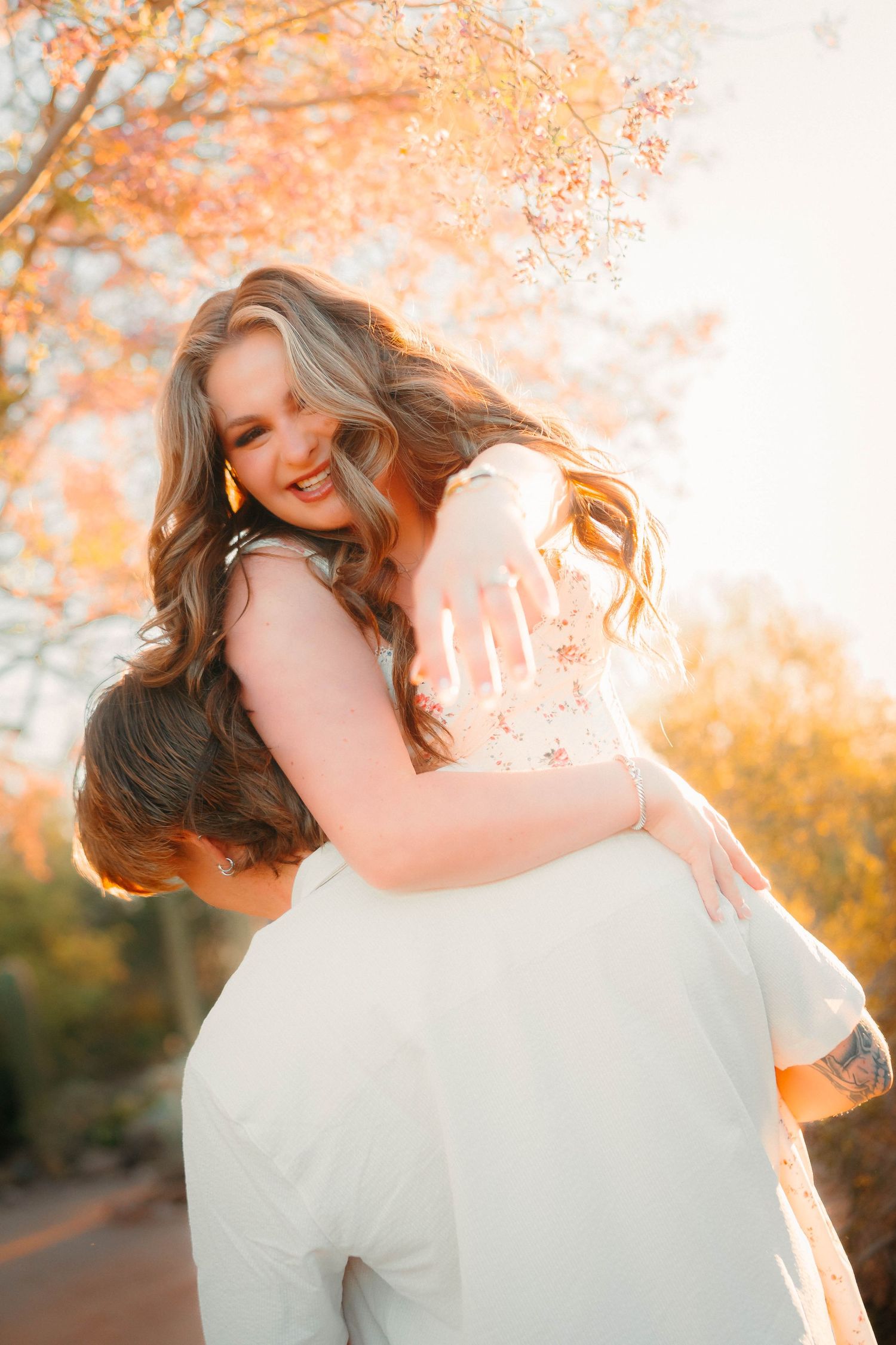 Couple embracing outdoors during golden hour, woman smiling joyfully while being lifted, autumn trees glowing in warm sunlight.