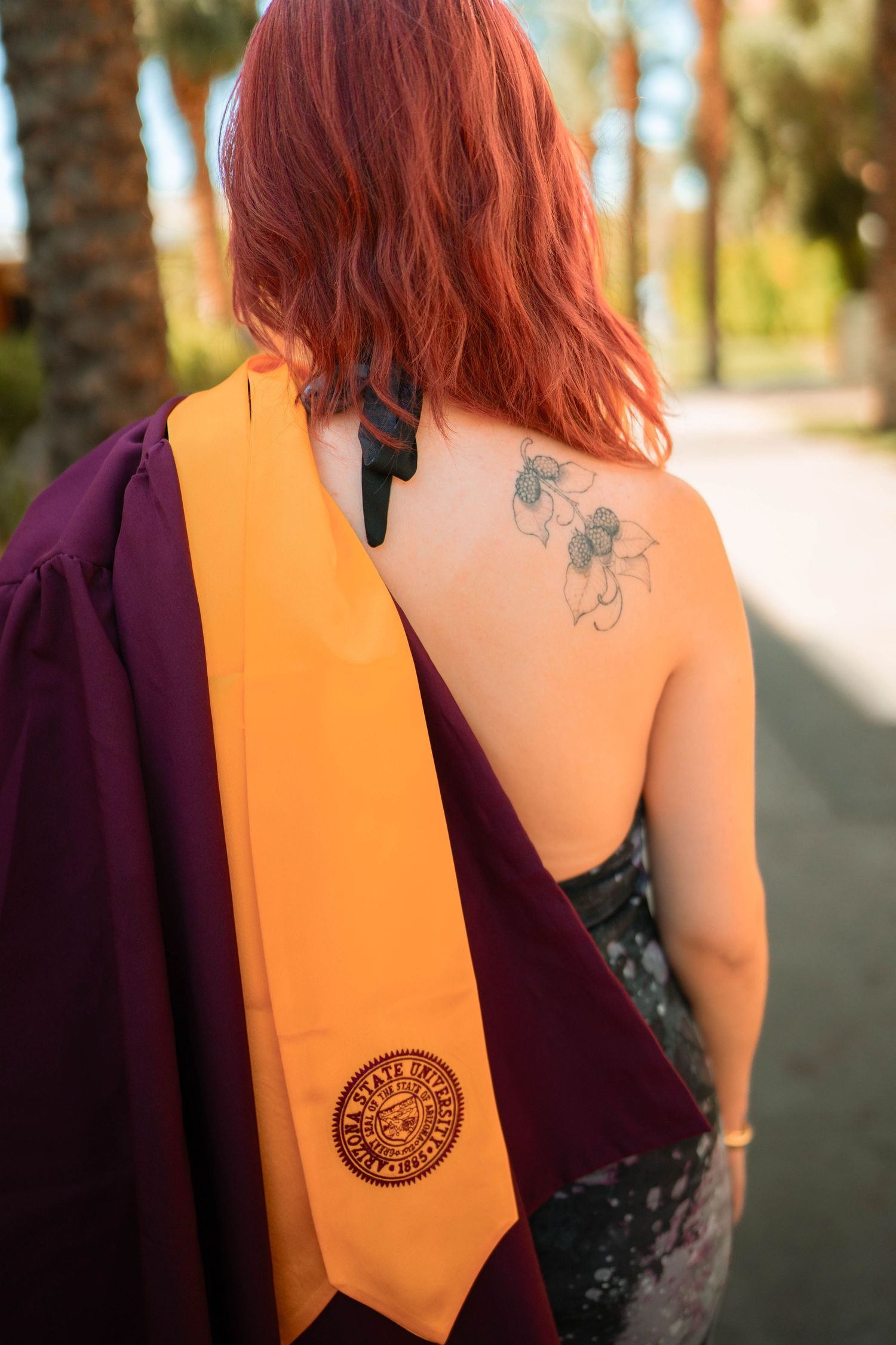 Red-haired graduate wearing maroon gown and gold ASU honor sash, showing floral back tattoo, outdoors near palm trees.