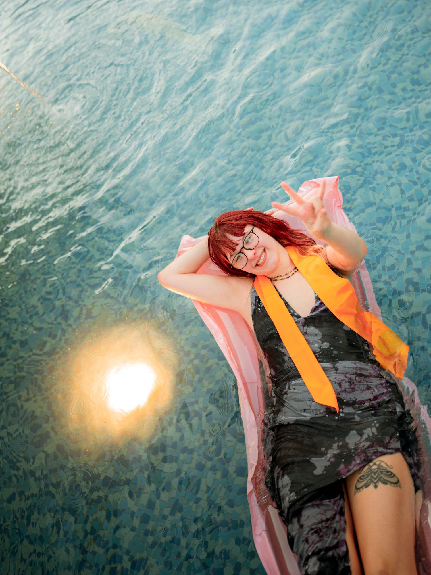 Woman with red hair and glasses relaxing by a pool, wearing a dark dress with an orange scarf.