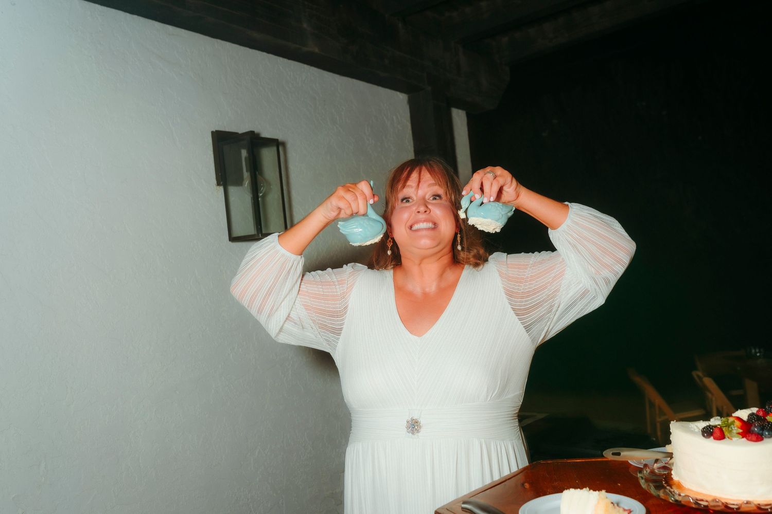 Bride in white lace dress playfully holding two pieces of wedding cake up to her ears and smiling.