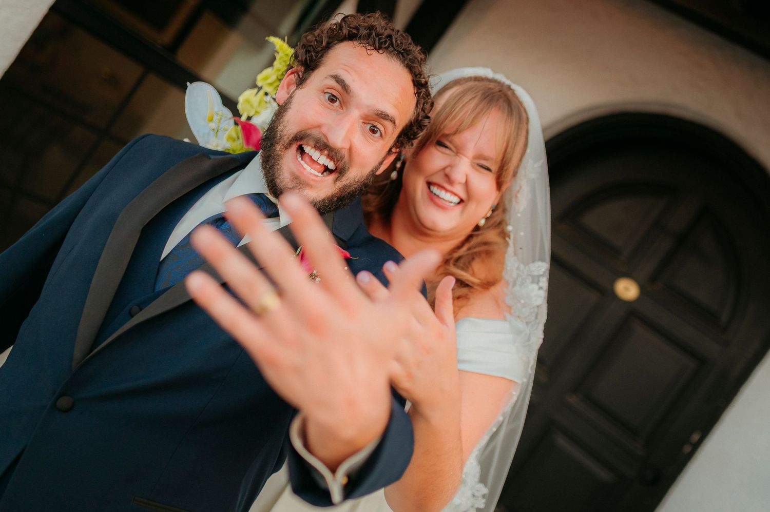 Smiling newlyweds showing off wedding rings on their fingers, bride in white veil and groom in blue suit.