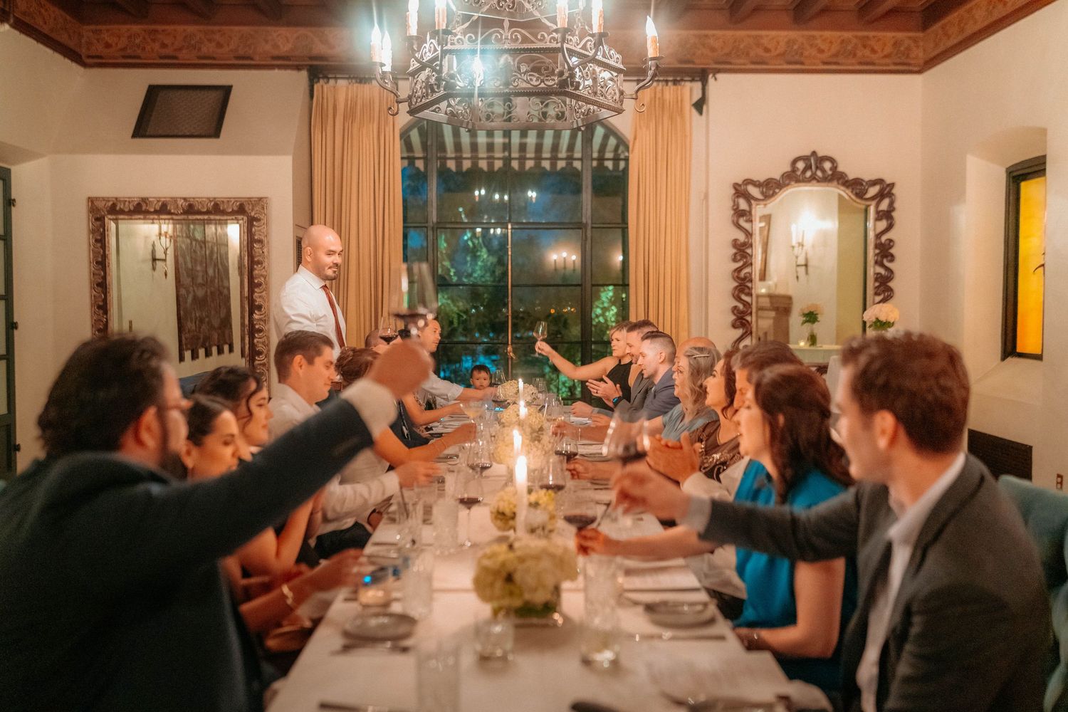 Guests raise glasses in a toast at an elegant wedding rehearsal dinner around a long candlelit table.
