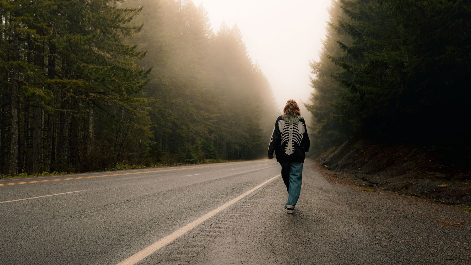 Woman walking down the center of a misty tree-lined road in a dark jacket and teal jeans.