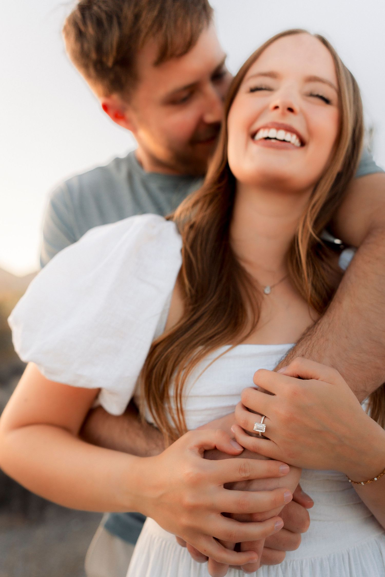 Couple embracing at sunset, woman laughing in white dress showing engagement ring.