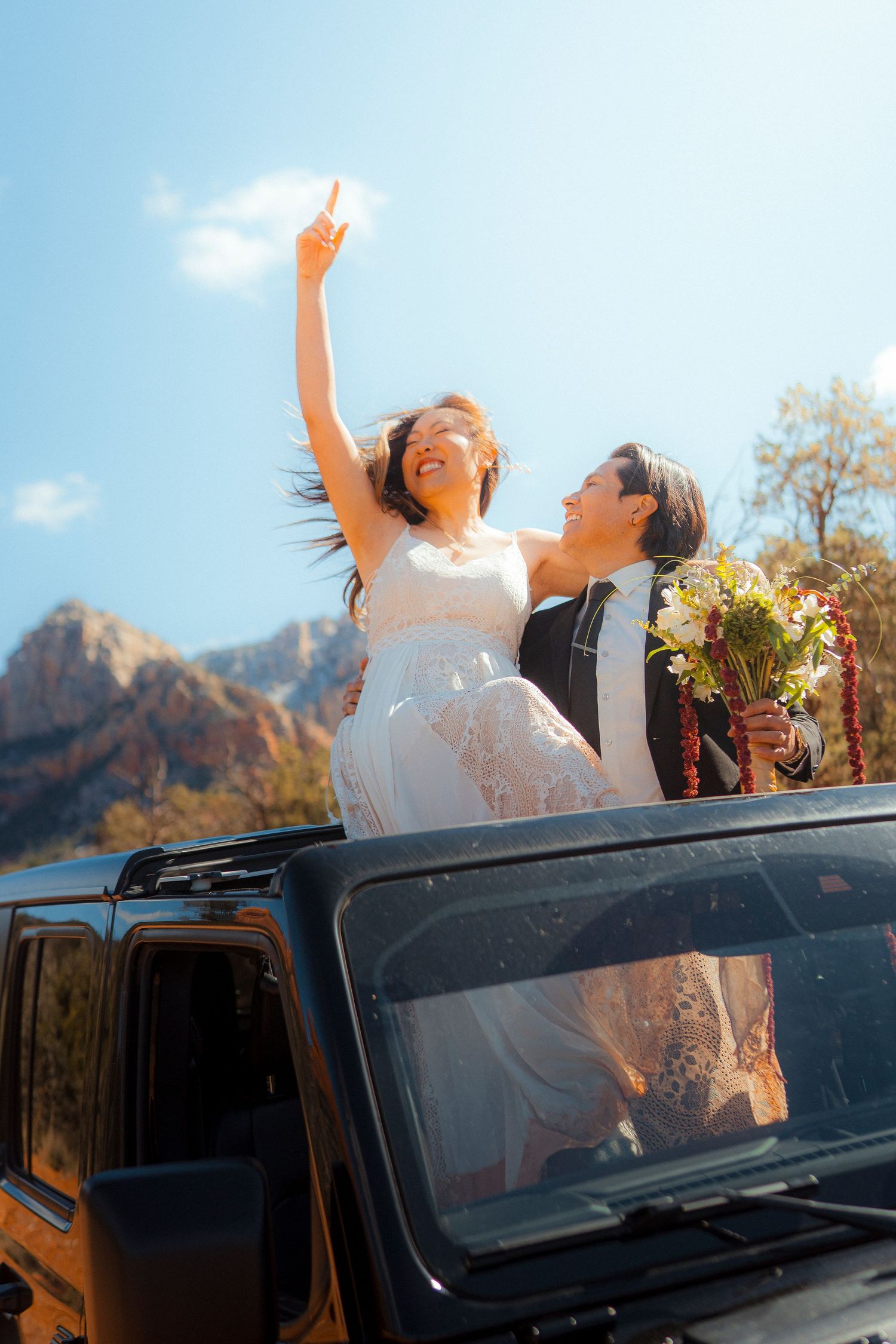 Bride and groom celebrate through sunroof of black Jeep with red rock mountains behind them.