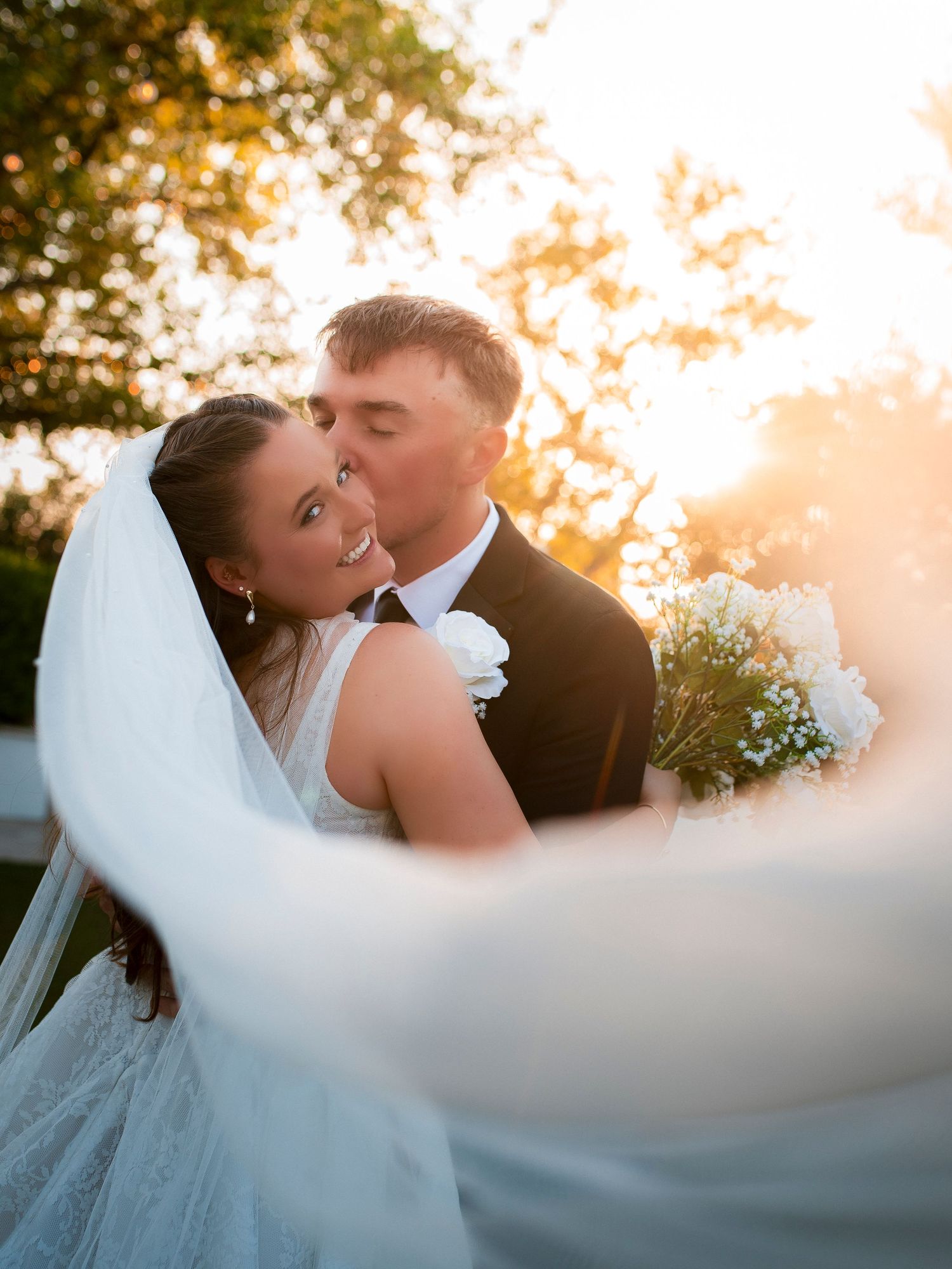 Groom kisses bride on the cheek in golden sunset light as her veil flows beautifully in the breeze.