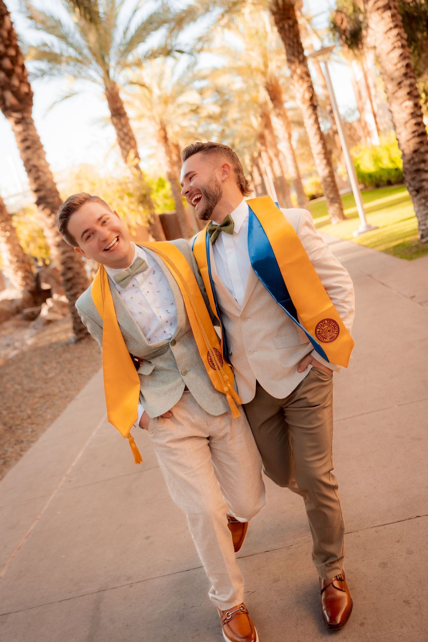 Two male graduates in suits with gold honor stoles laughing while walking under palm trees.