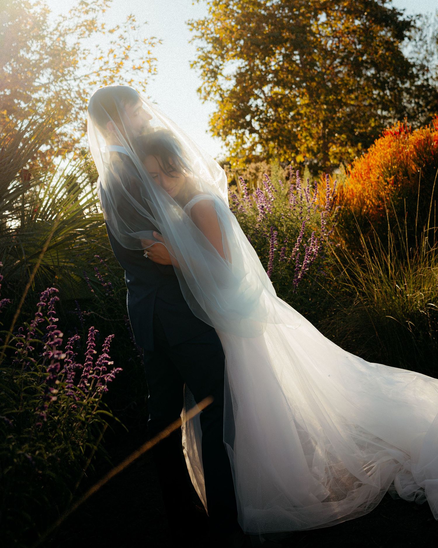 Bride and groom kissing under a veil in a sunlit garden with colorful wildflowers and autumn trees.
