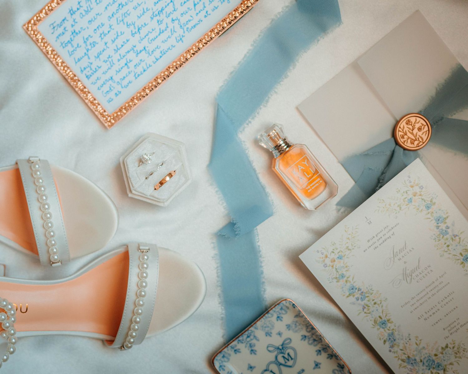 Elegant wedding flat lay featuring blue heels, perfume, floral stationery, and pearl accessories on a light blue background.