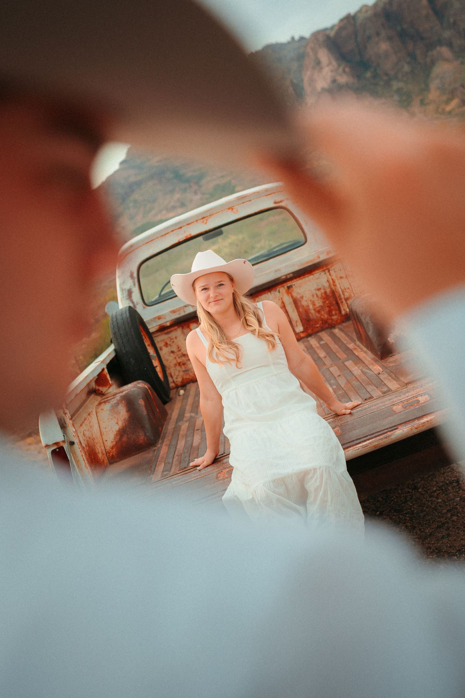Woman in white dress and cowboy hat leaning against rusty vintage truck with mountains in background.