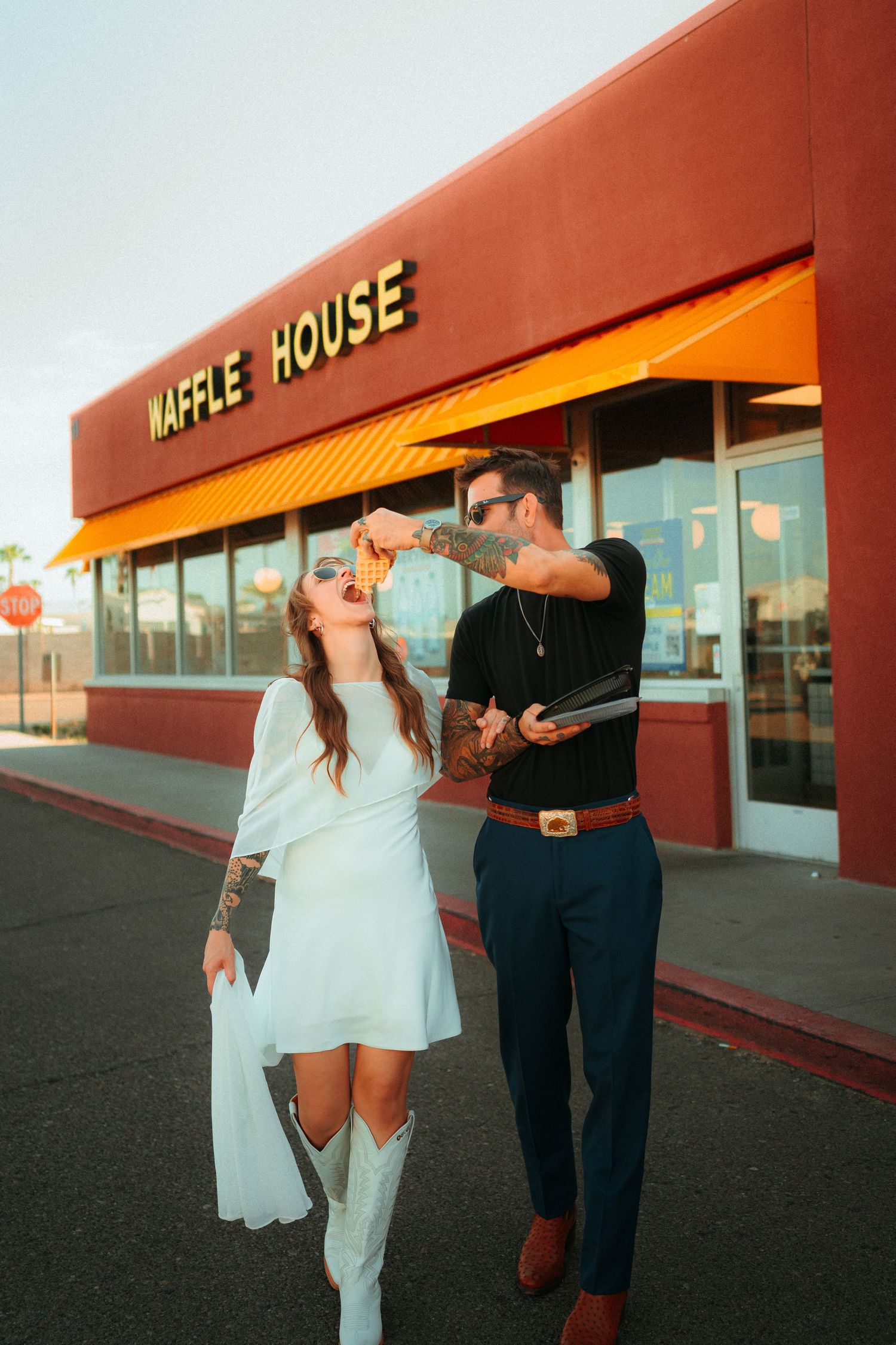 Couple in wedding attire sharing food outside Waffle House restaurant, woman in white dress and boots, man in black shirt.