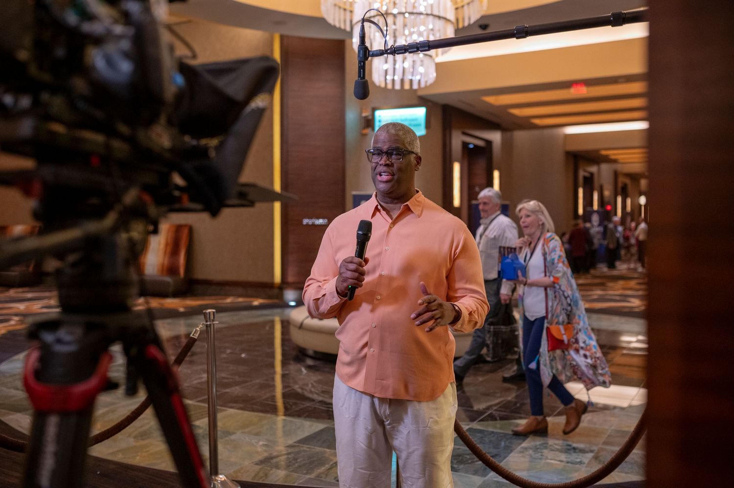 Man in pink shirt speaks into microphone on camera at indoor event.