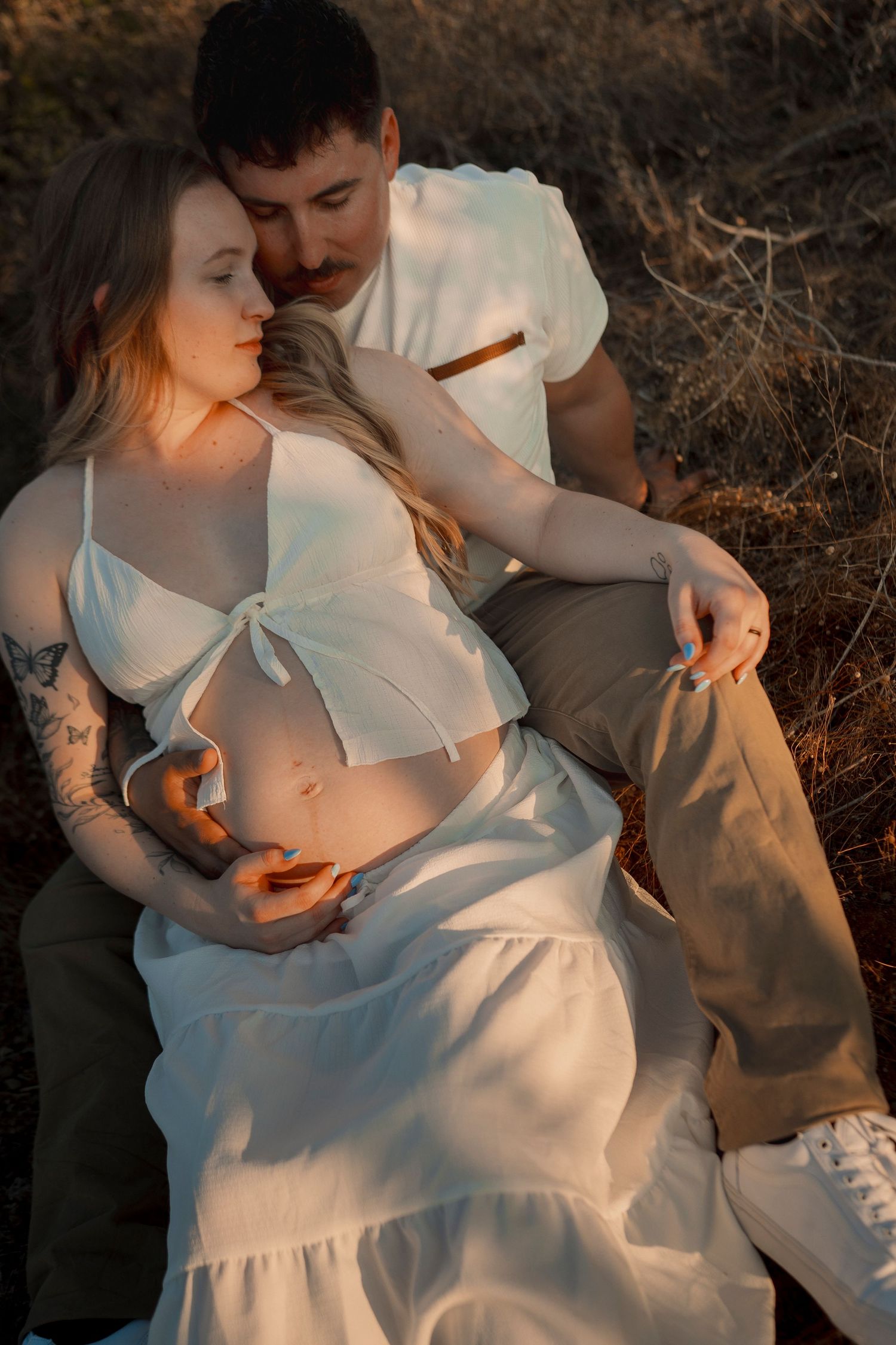 Pregnant woman in white dress cradled by partner in golden sunset light during maternity photo shoot.