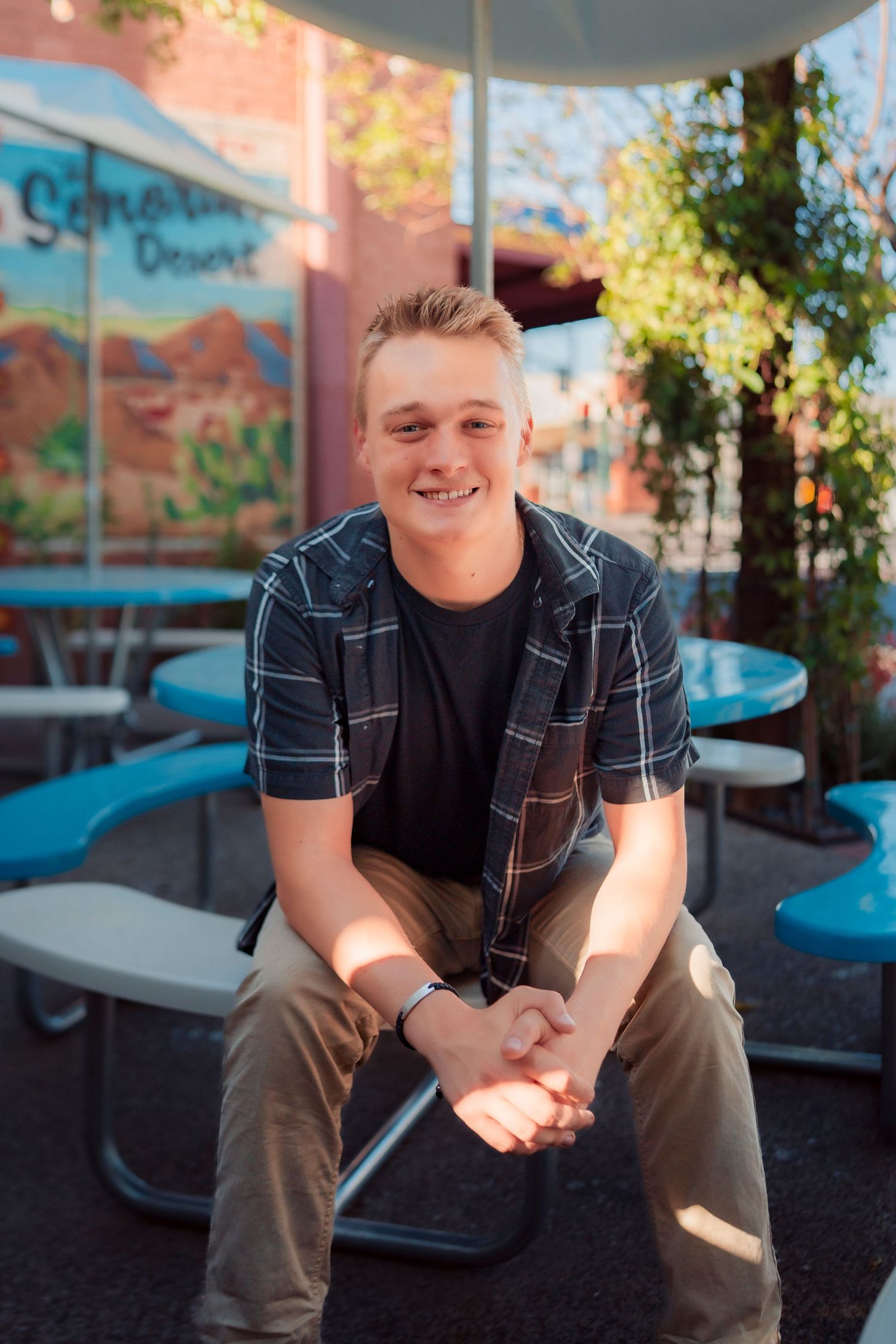 Smiling young man in plaid shirt sits at outdoor patio table in sunny urban setting.