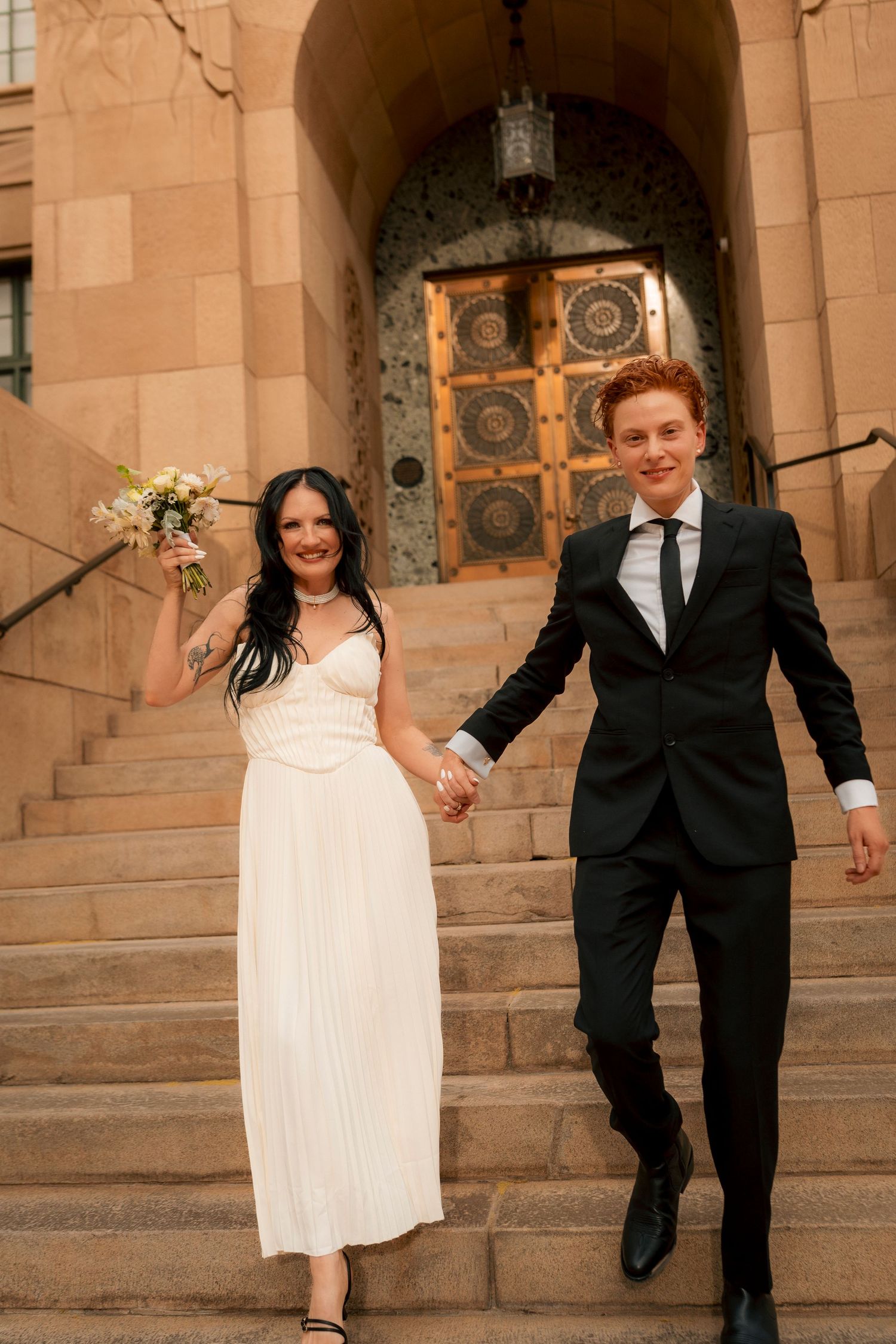 Newlywed couple descending stone church steps, bride in white dress holding bouquet, groom in black suit, holding hands.