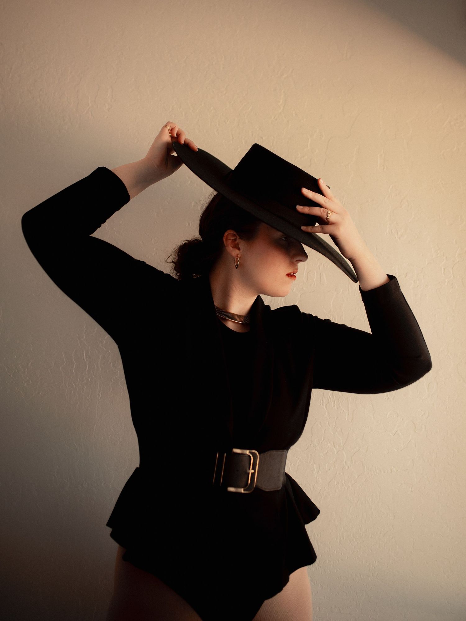 Woman in black bodysuit and wide-brim hat poses dramatically against a neutral wall with a wide belt accent.