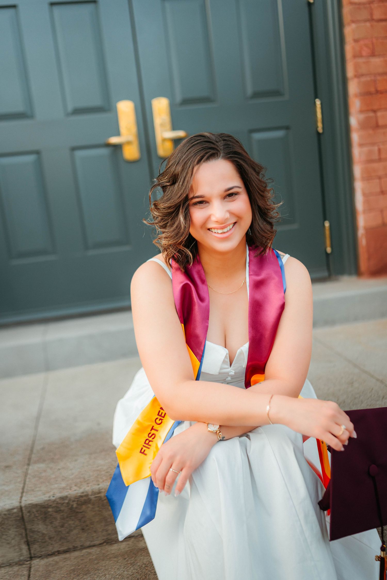 Smiling graduate in white dress wearing colorful sashes sits on steps outdoors.