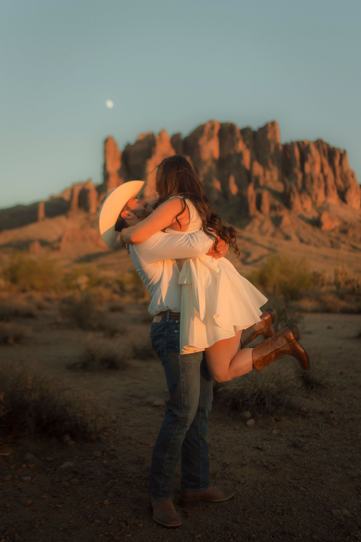 Man in cowboy hat lifting woman in white dress in a desert landscape with rocky mountains at dusk.