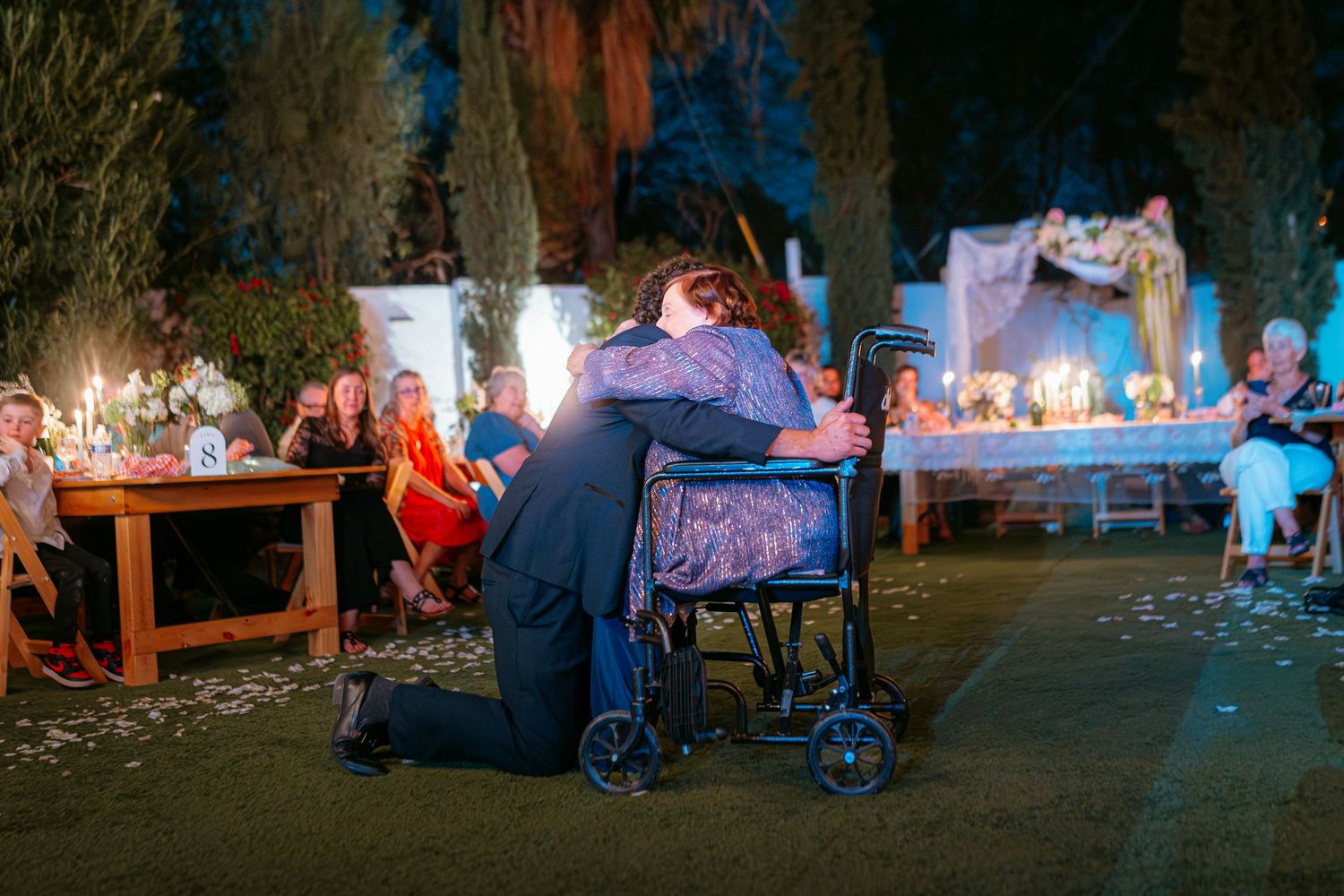 Person kneeling to embrace someone in a wheelchair at an outdoor evening wedding reception with candles.