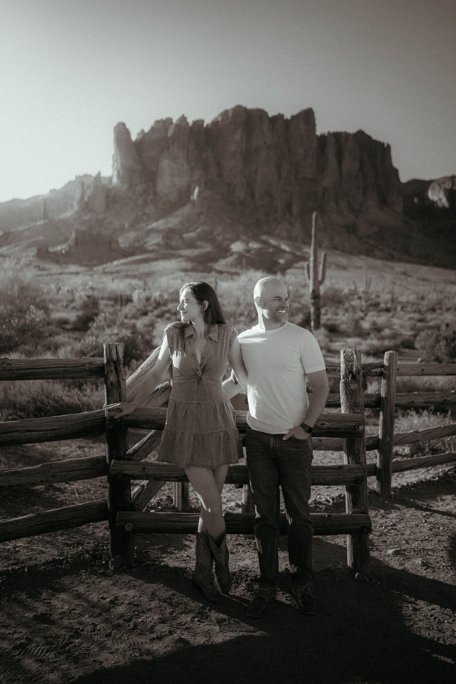 Black and white photo of couple leaning on rustic fence with majestic Superstition Mountains behind them.