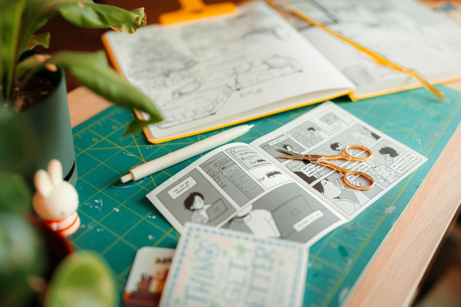 Comic zine pages with scissors on a green cutting mat beside a Miffy figurine.
