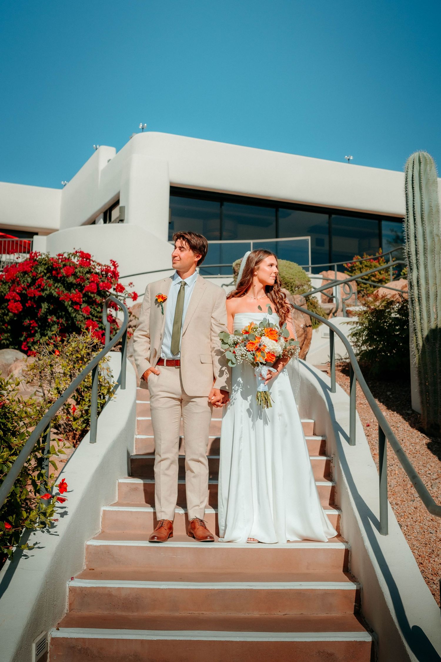Bride and groom hold hands on outdoor staircase with cacti and red flowers at Scottsdale desert wedding venue.