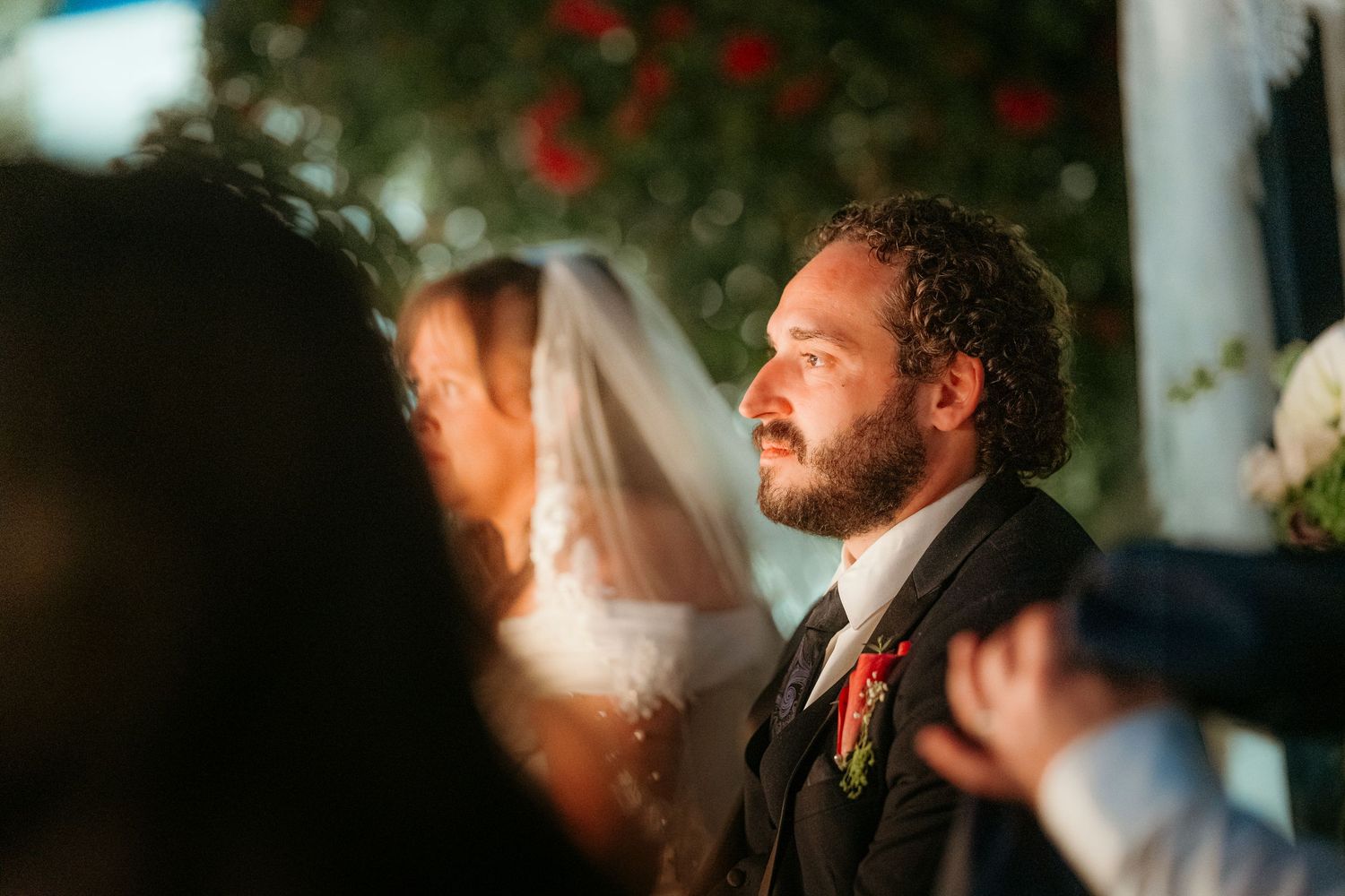 Groom in black suit gazing emotionally during outdoor wedding ceremony with bride in veil in background.
