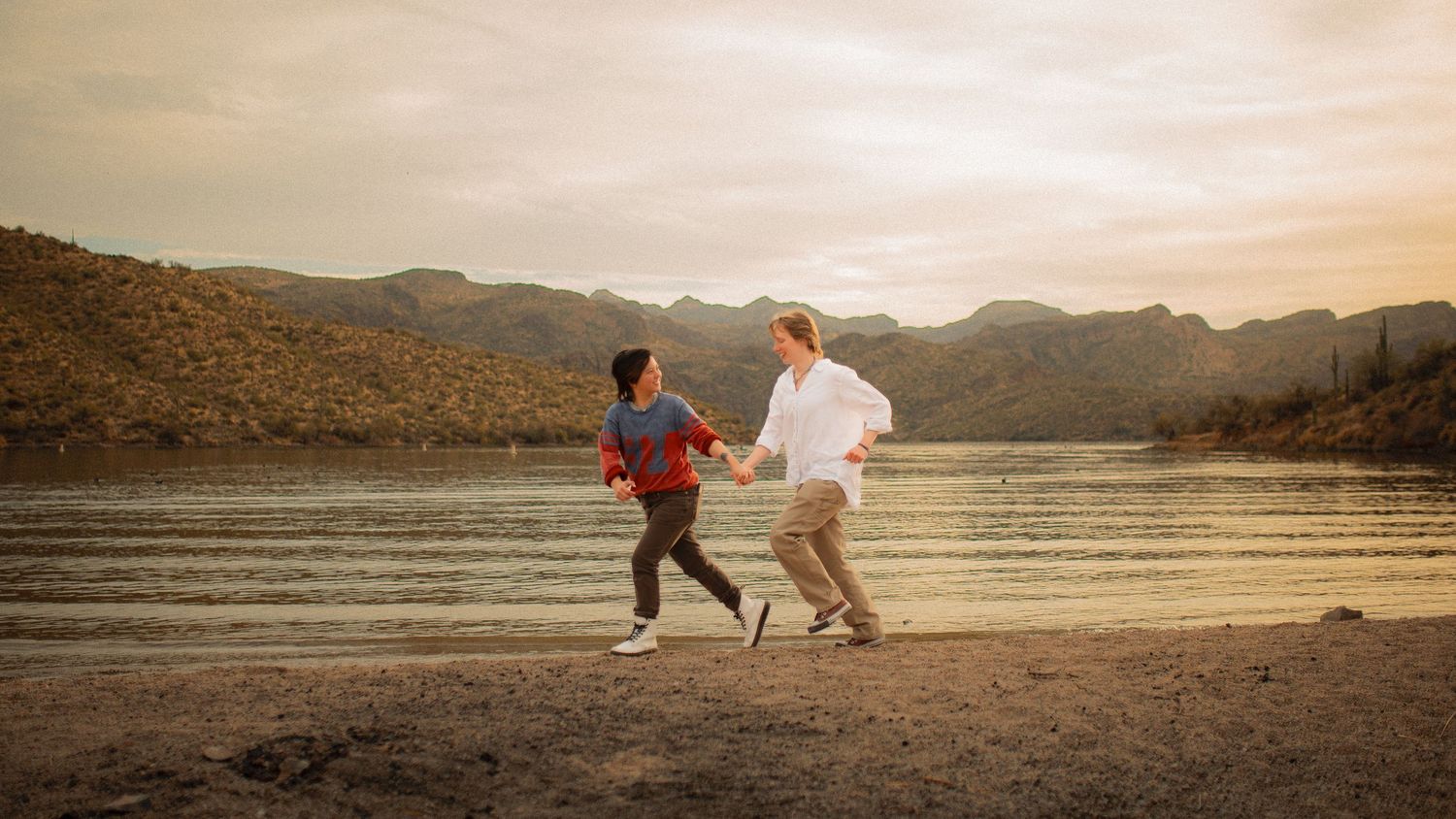 Couple running and laughing along a scenic lakeside beach at golden sunset with mountain backdrop.