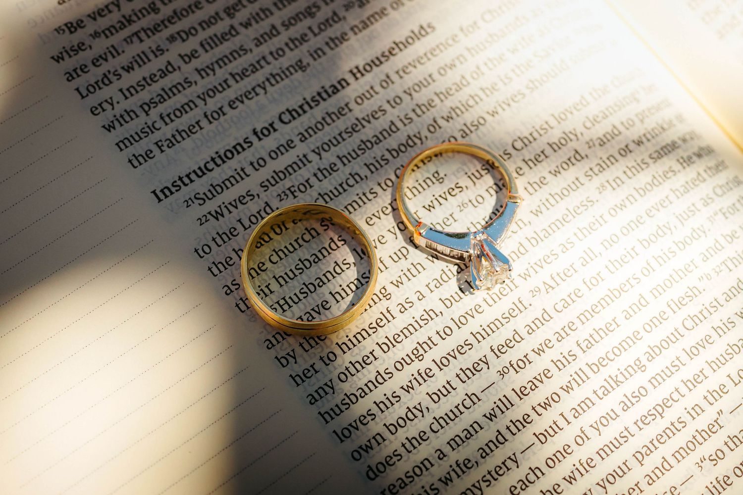 Two gold wedding rings rest on an open Bible page showing Christian marriage instructions in soft light.