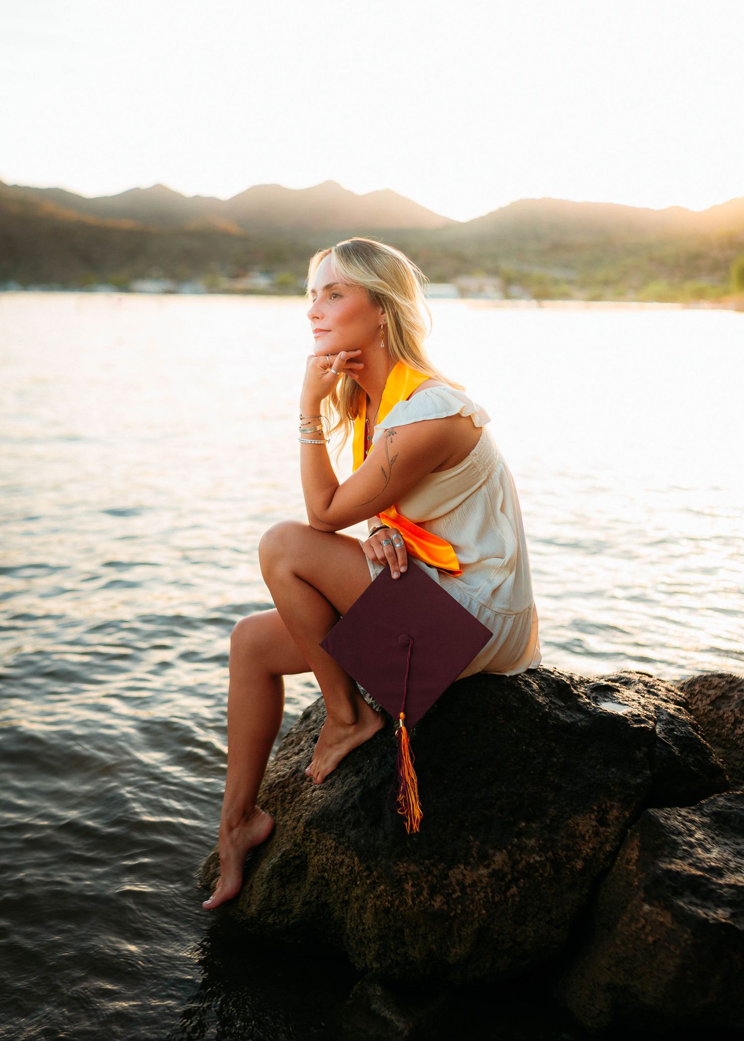 Blonde graduate sitting on rocks by ocean at golden sunset, holding maroon cap with orange tassel.
