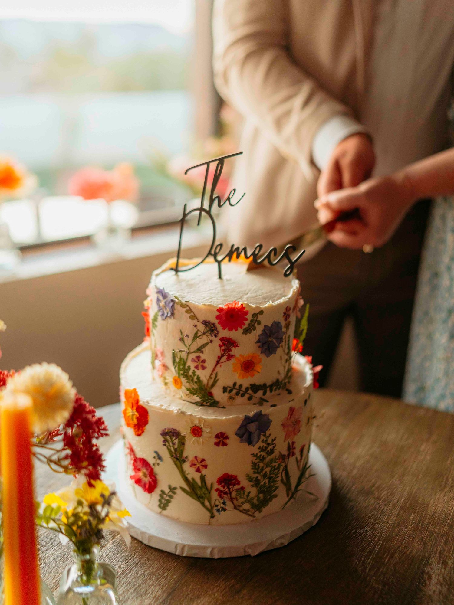 Two-tier wedding cake decorated with colorful pressed flowers and The Demees cake topper.