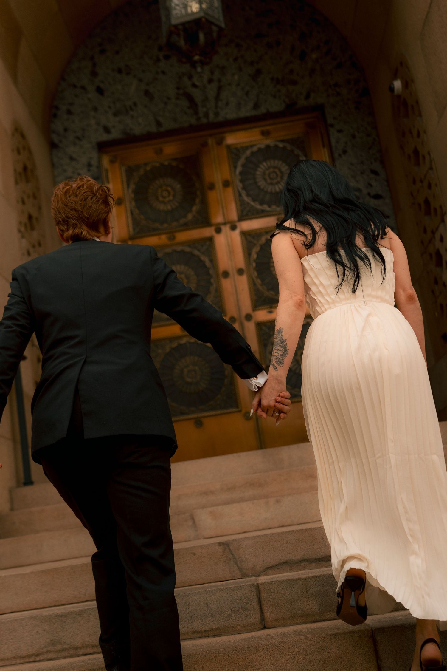 Bride in white dress and groom in black suit hold hands walking up stone steps toward ornate golden doors.