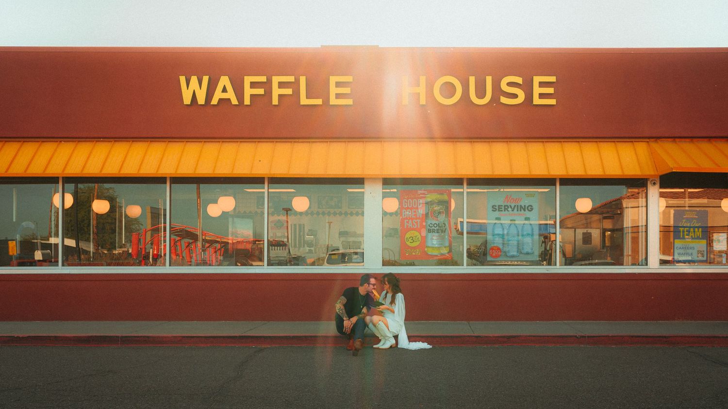 Couple sitting together in front of a Waffle House restaurant with bright red and yellow signage at sunset.
