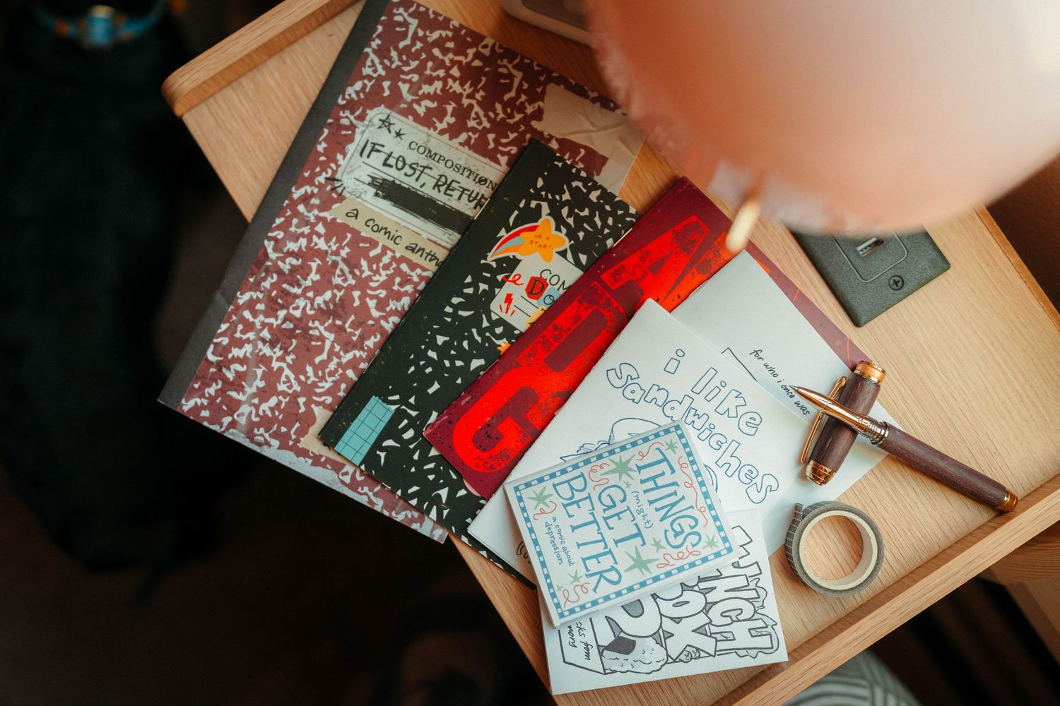 Composition notebooks, stickers, pen, and washi tape arranged on a wooden nightstand.
