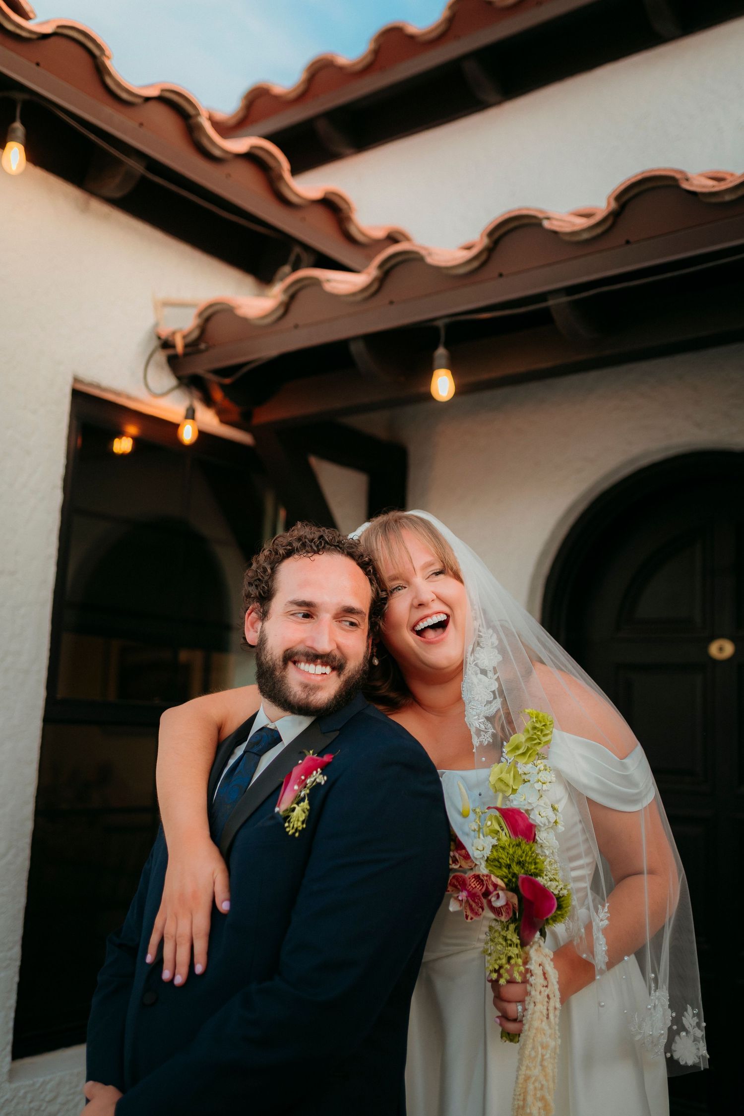 Laughing bride in white gown embracing groom in navy suit outside Spanish-style venue.