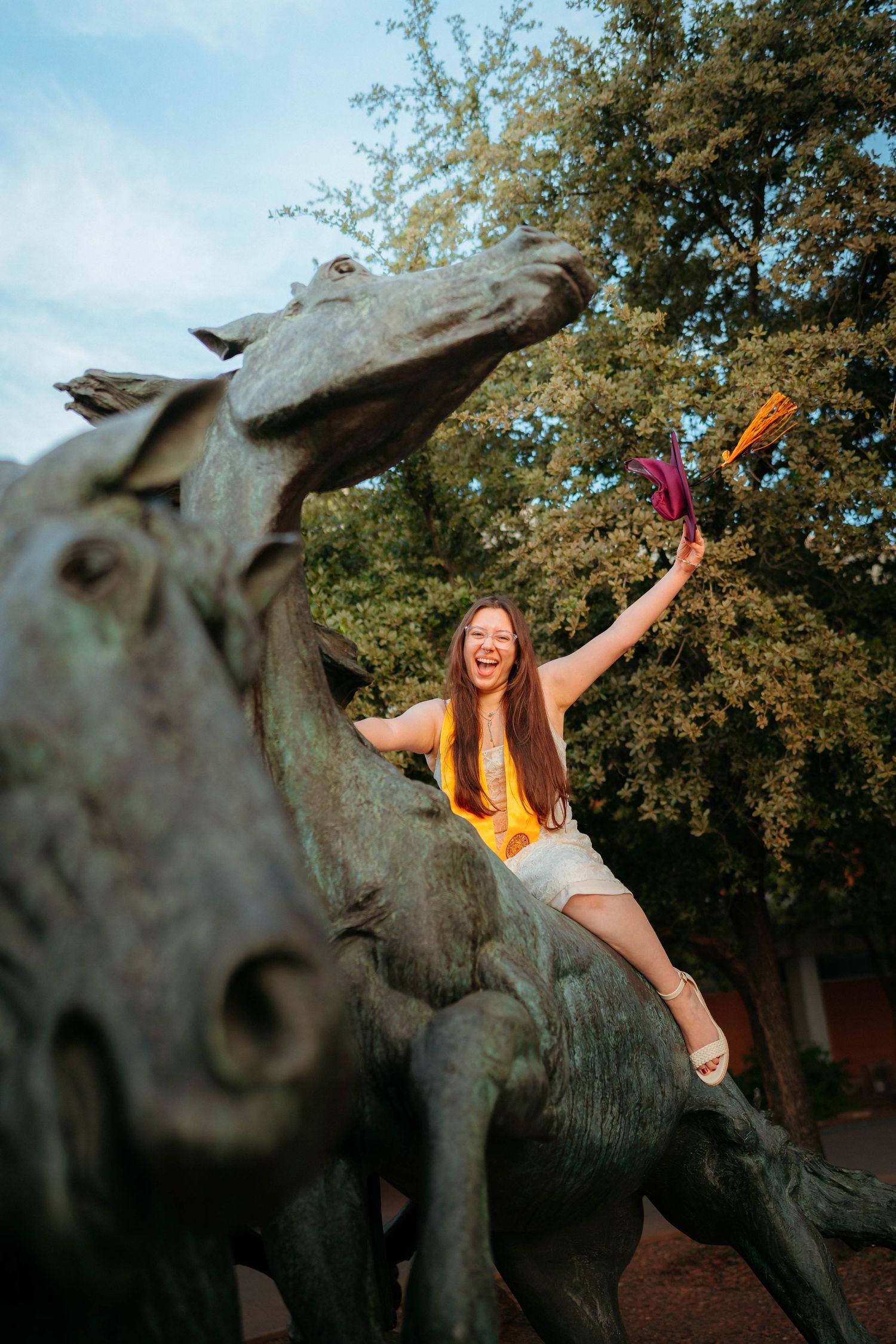 Excited graduate in yellow sash sits on bronze horse statue outdoors holding colorful tassels.