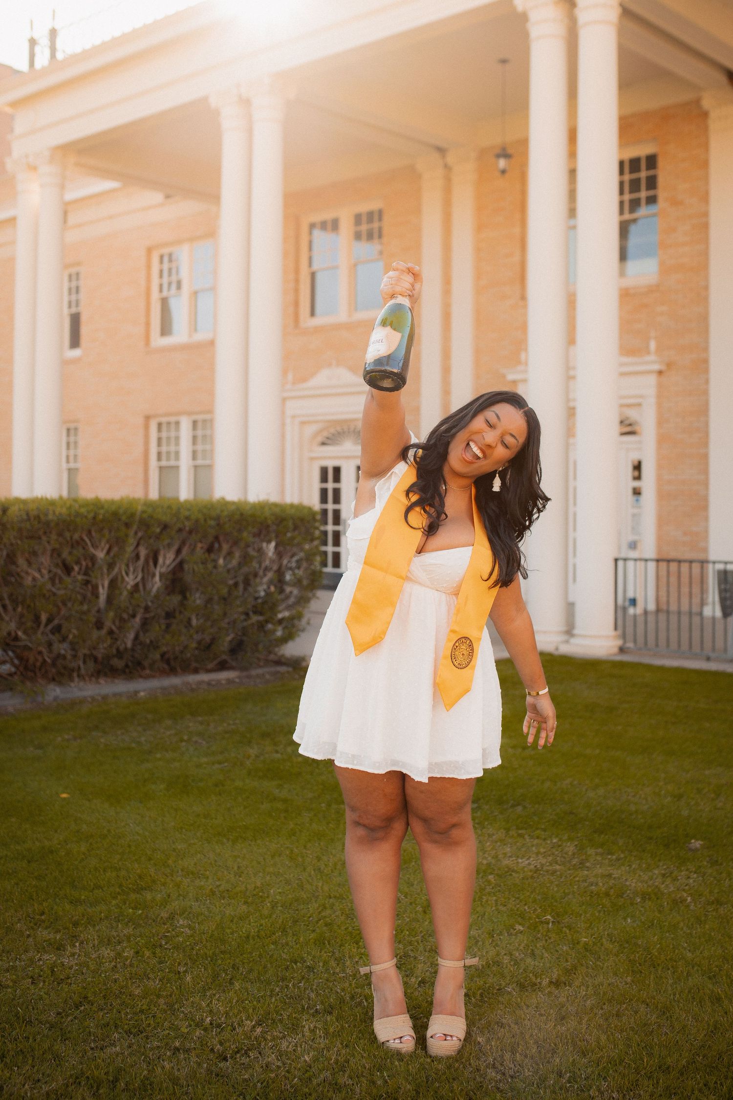 A graduate in white dress raises a champagne bottle wearing a gold ASU stole.