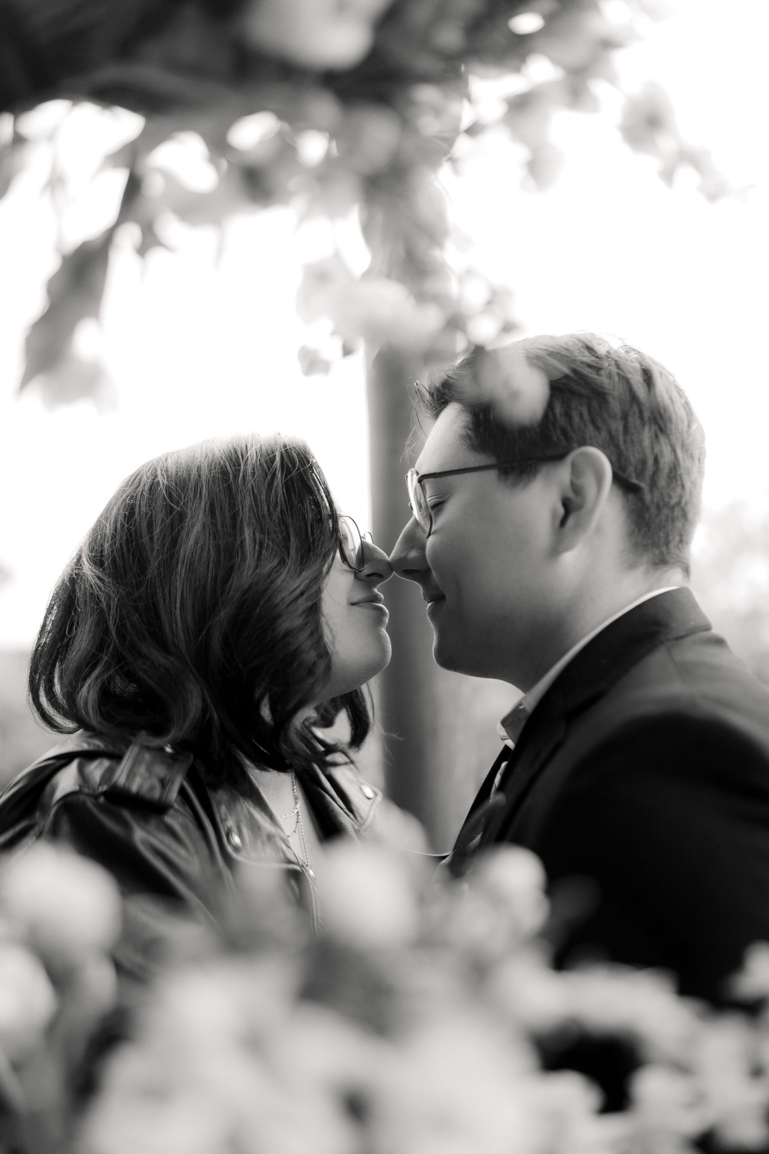 Black and white romantic close-up of couple touching noses under floral canopy.