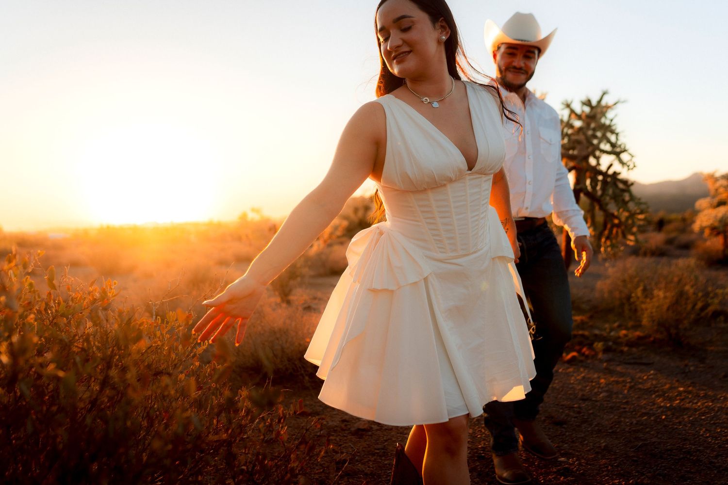 Bride in short white dress and groom in cowboy hat walk through golden desert field at sunset.