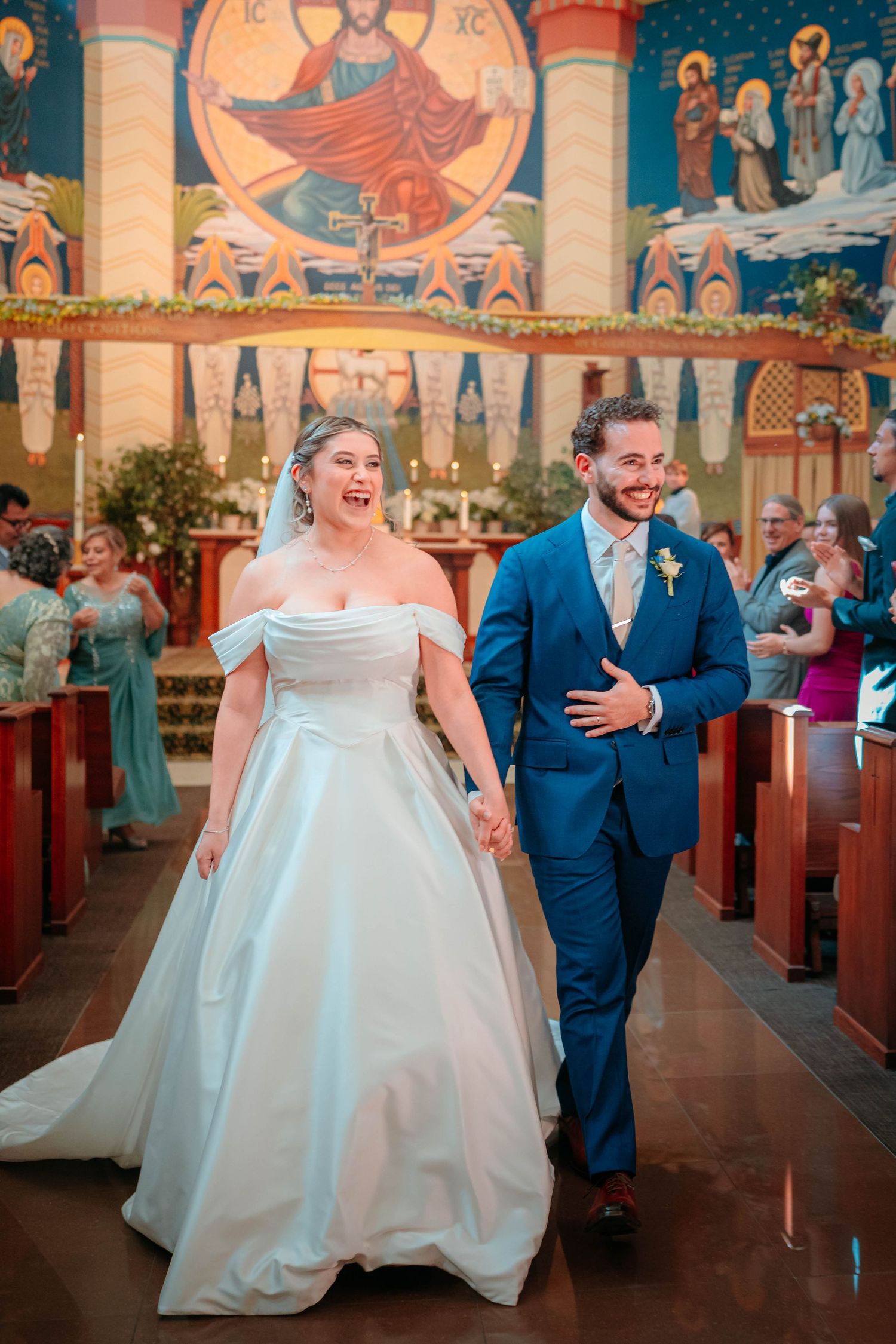 Newlyweds joyfully walking down church aisle after ceremony in ornate Byzantine-style decorated chapel.