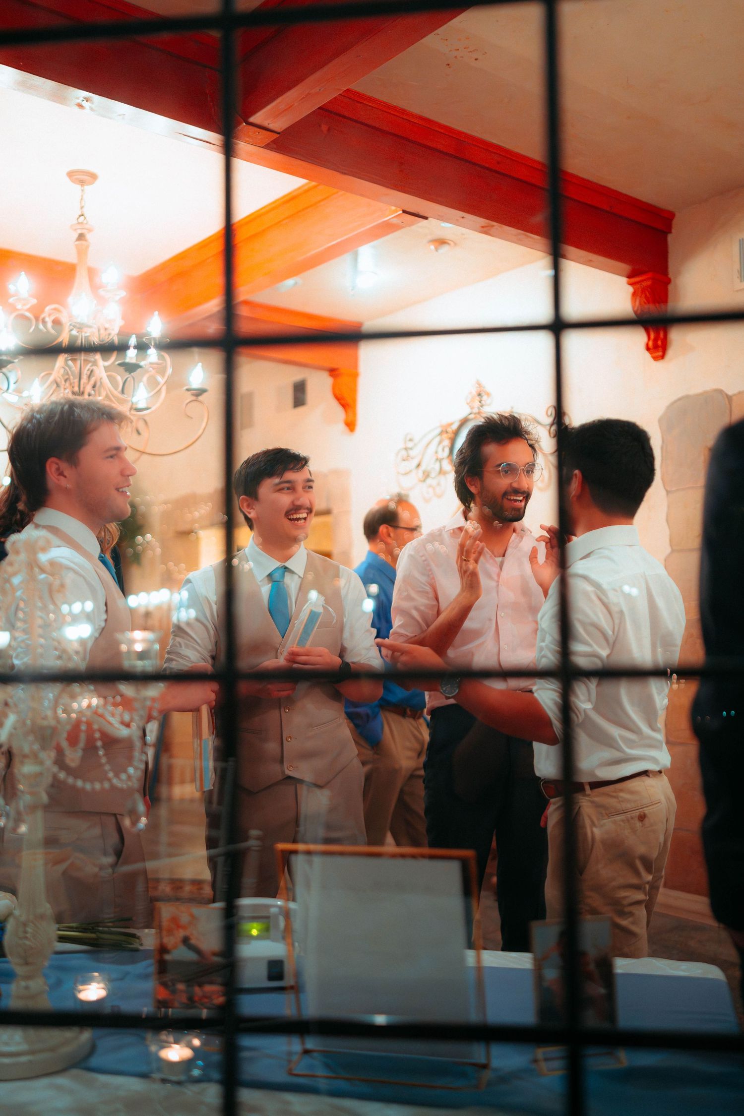 Men in dress shirts laughing and socializing at an indoor gathering, viewed through a window with chandeliers overhead.