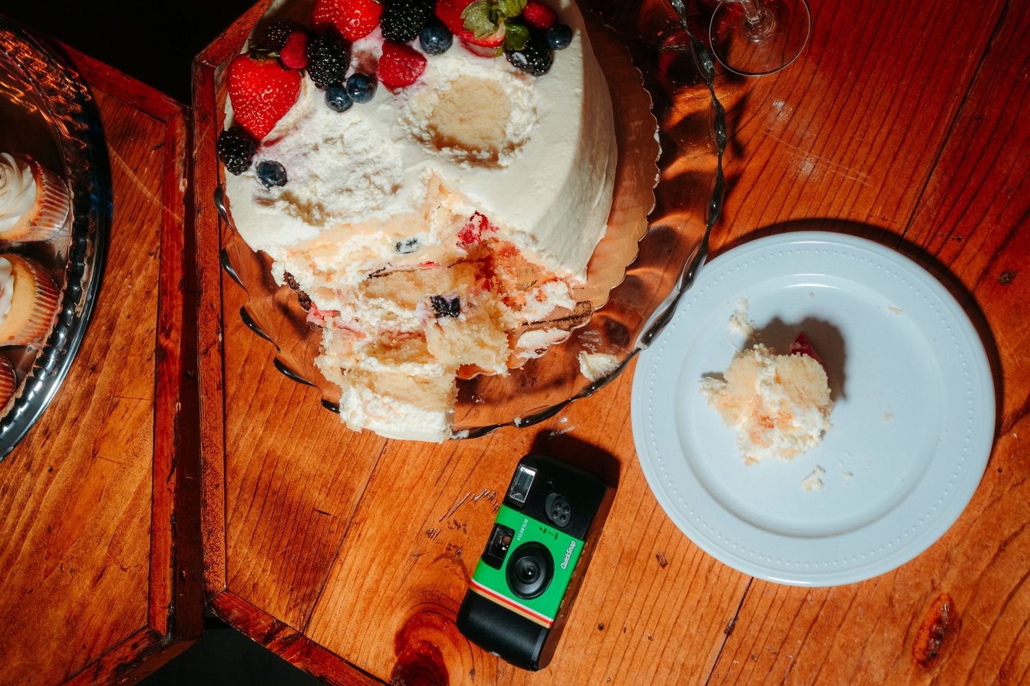 A partially sliced white wedding cake topped with fresh berries sits on a wooden table beside a dessert plate.