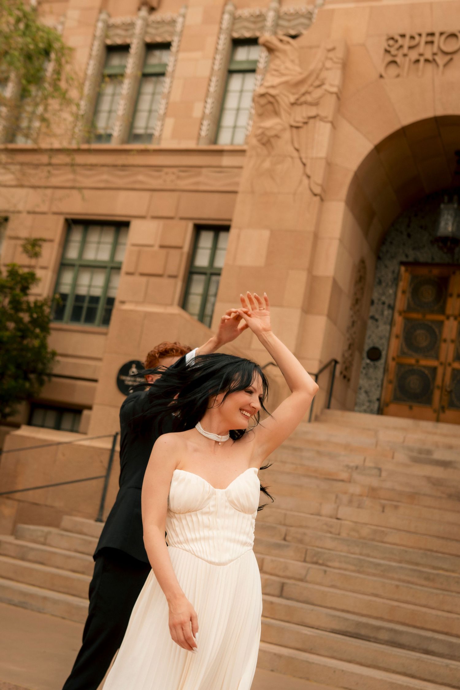 Bride in white gown twirls with groom in black suit on steps of ornate stone building.