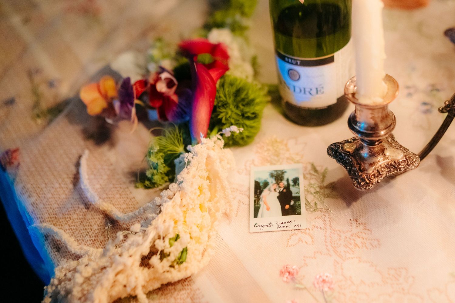 A wedding table display with a Polaroid photo, champagne bottle, candle holder, and colorful floral arrangement on a lace cloth.
