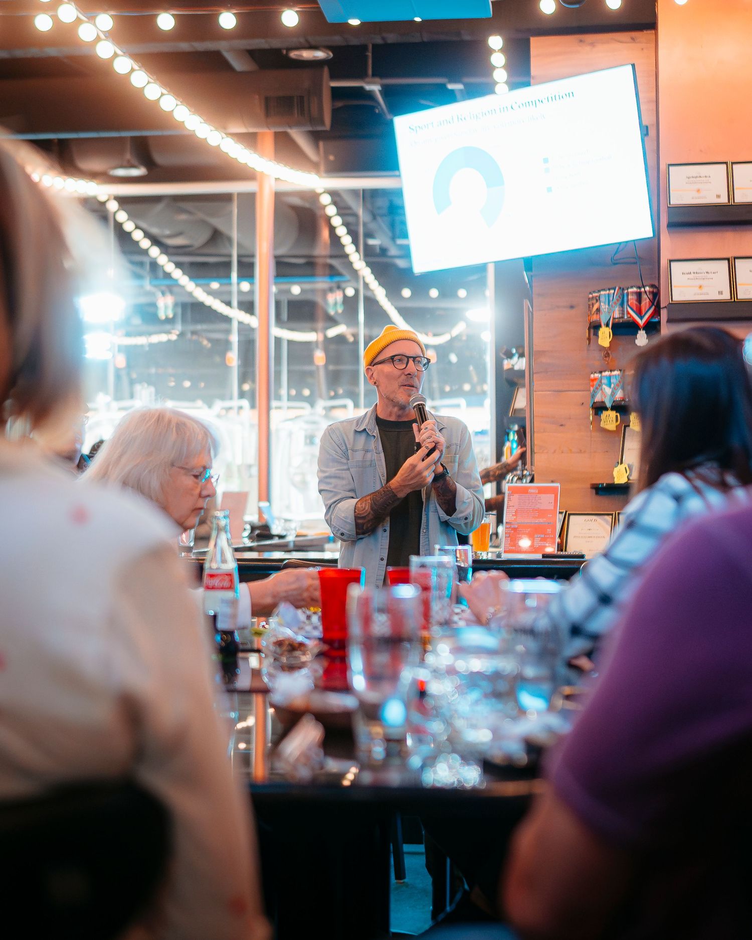 Man in yellow hat speaking to a crowd at a brewery event with string lights.