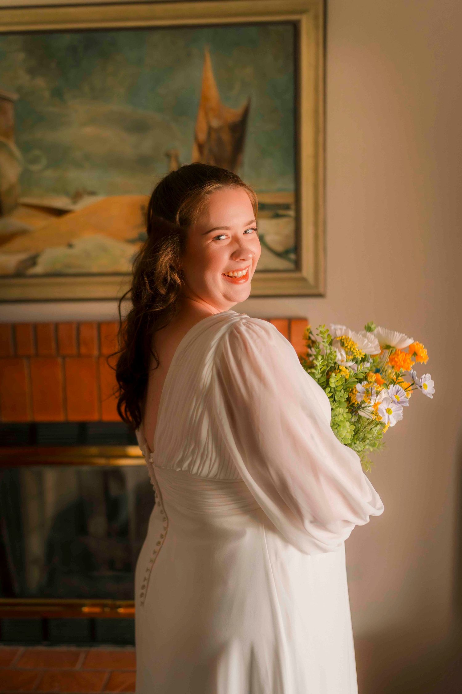 Smiling bride in white gown holding yellow and white bouquet beside a brick fireplace with artwork.