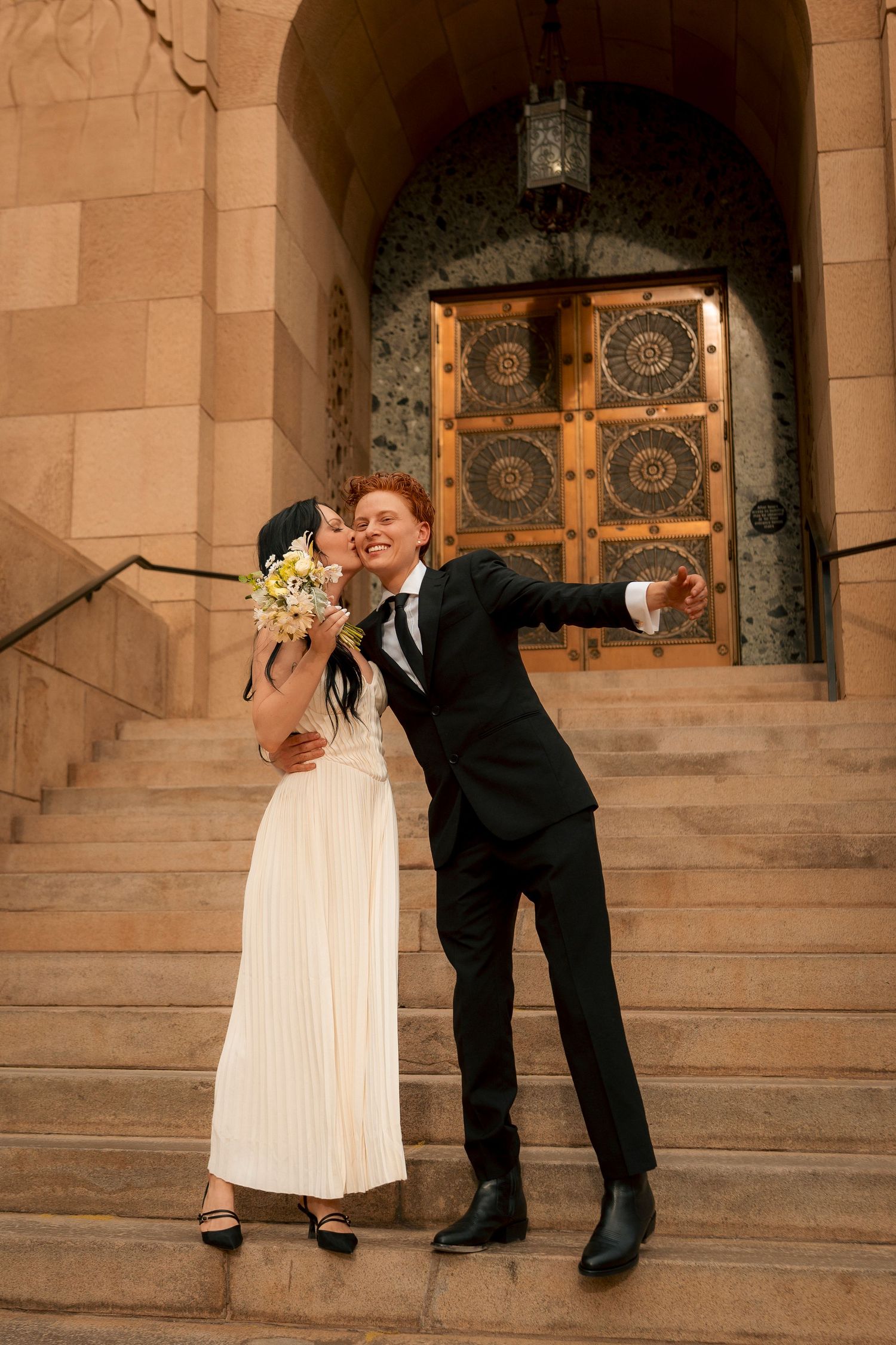 Bride in white dress holding bouquet embraces groom in black suit on stone church steps with ornate doors.