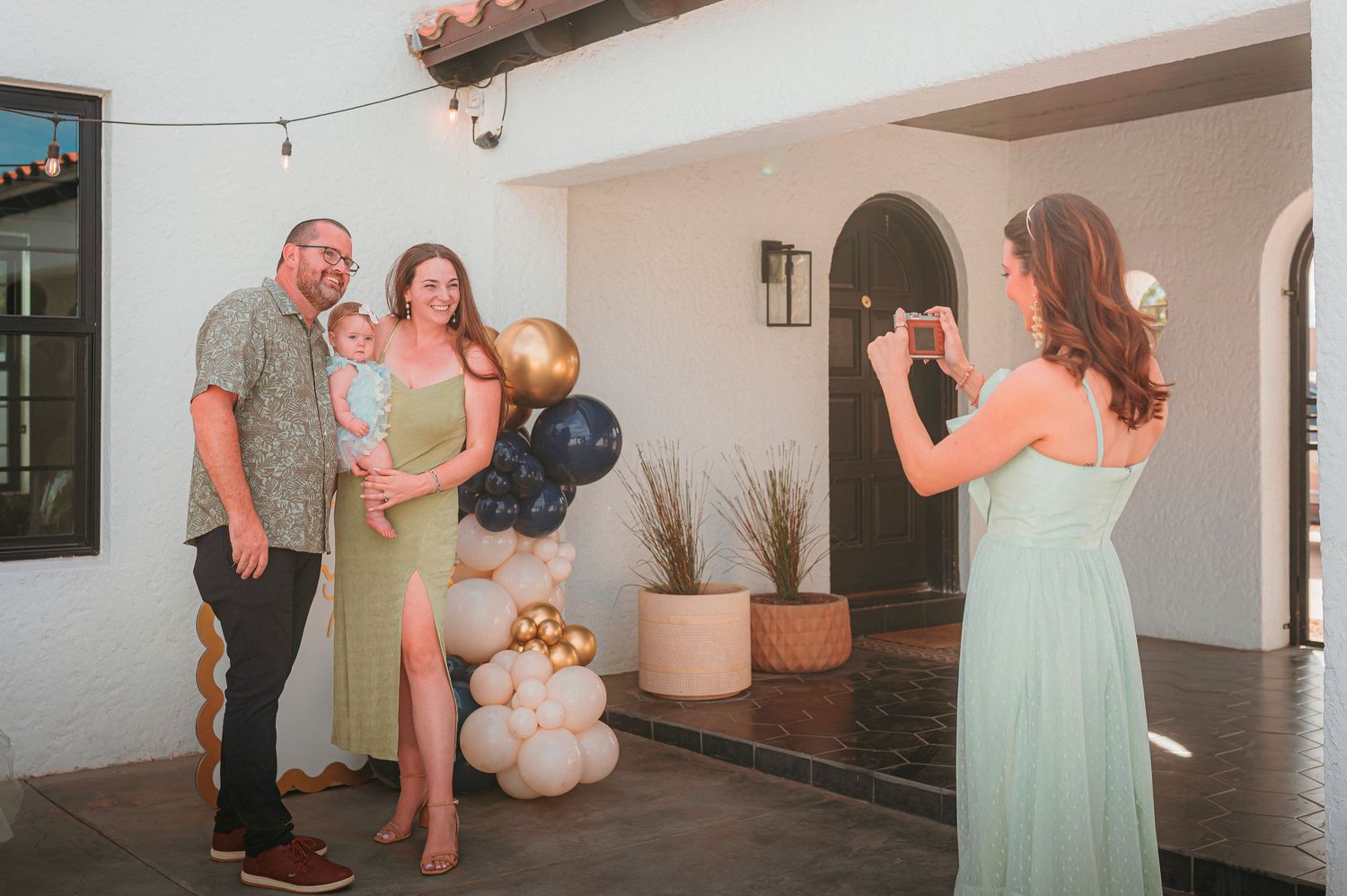 A woman photographs a couple holding a baby near gold, black, and white balloons at an outdoor celebration.